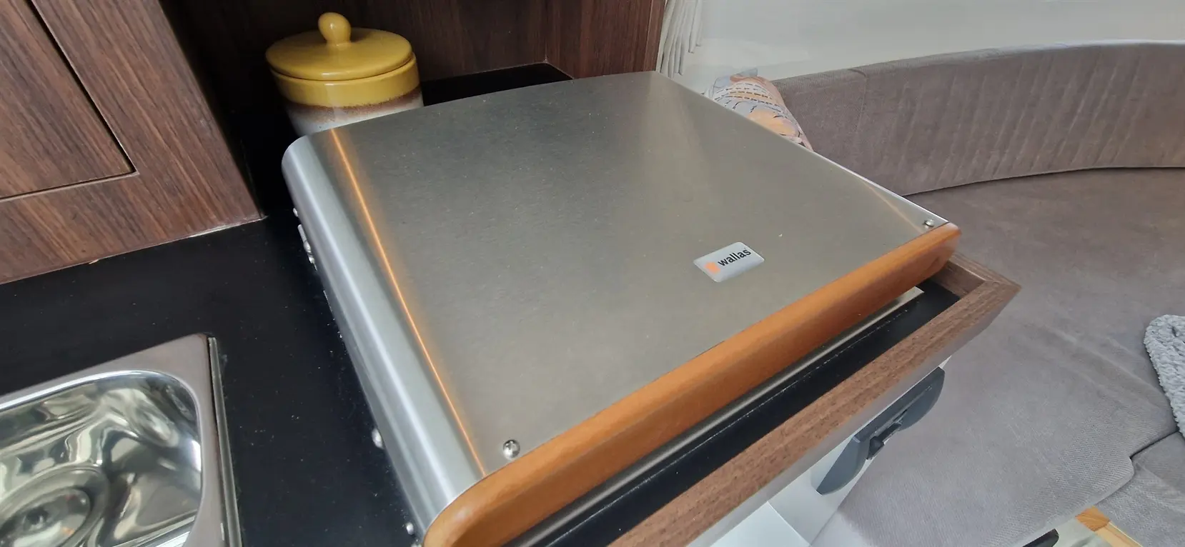 A metallic cooking surface with wooden trim on a dark countertop, alongside a yellow lidded container and a sofa.